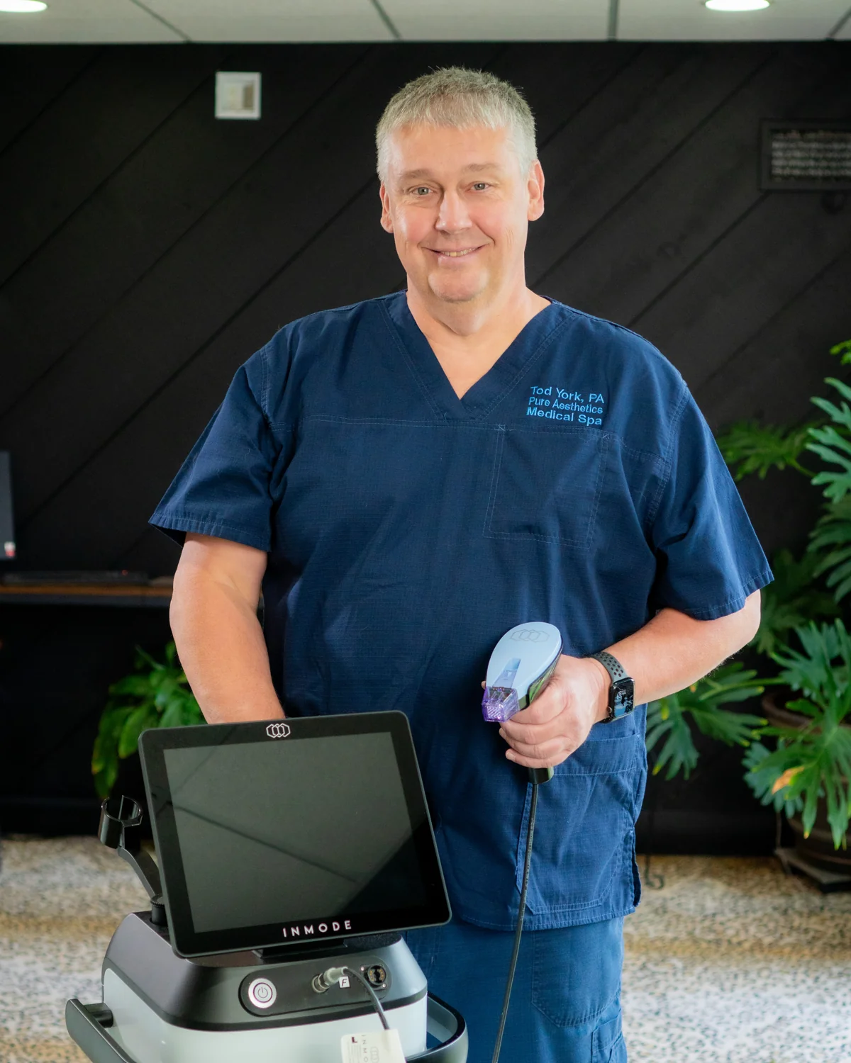 Tod York, PA, board-certified physician assistant, in navy scrubs beside InMode aesthetic equipment